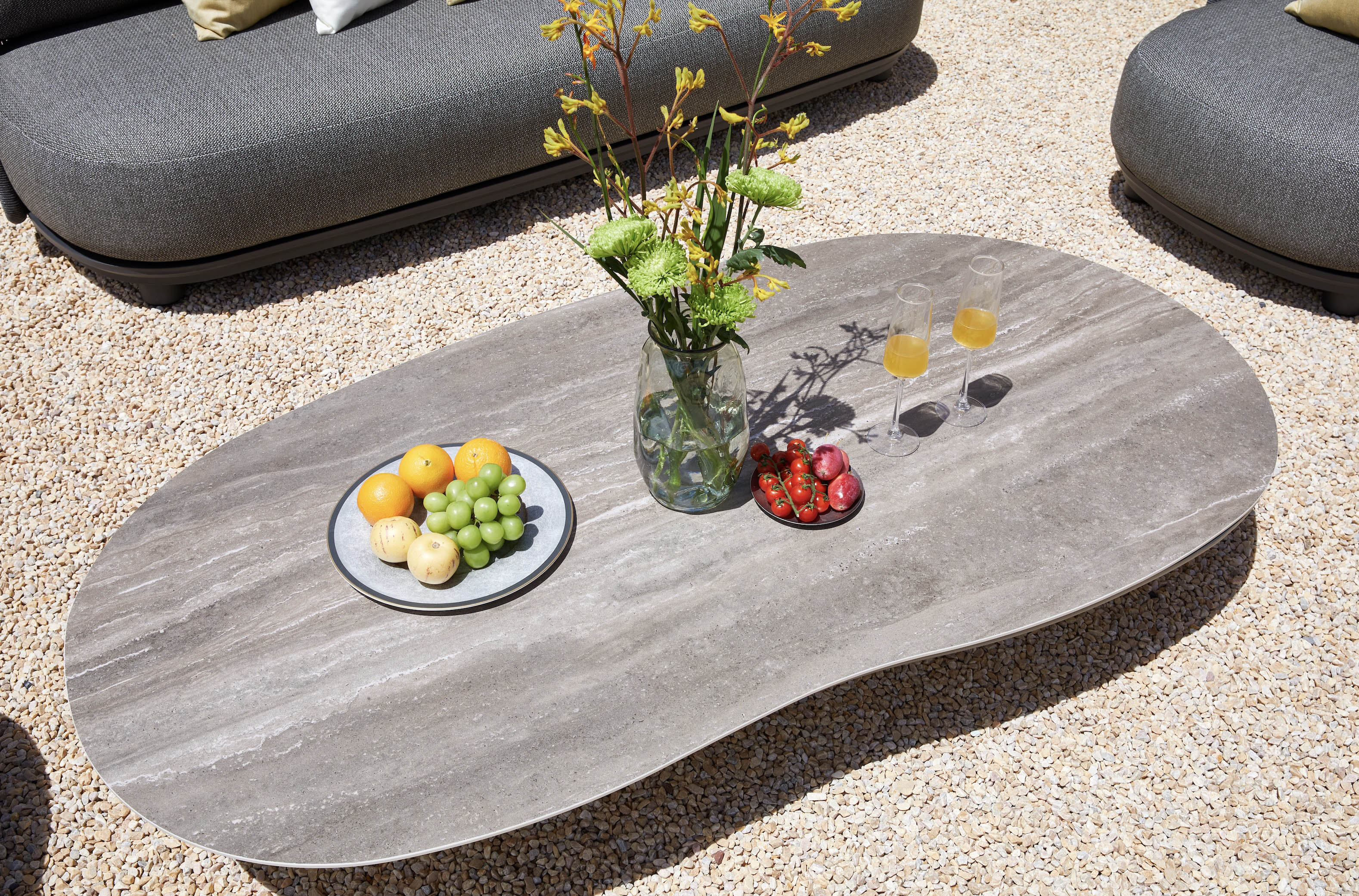 Table basse ovale en aluminium design avec plateau céramique gris effet pierre, décorée avec vase et fruits, ambiance luxe.