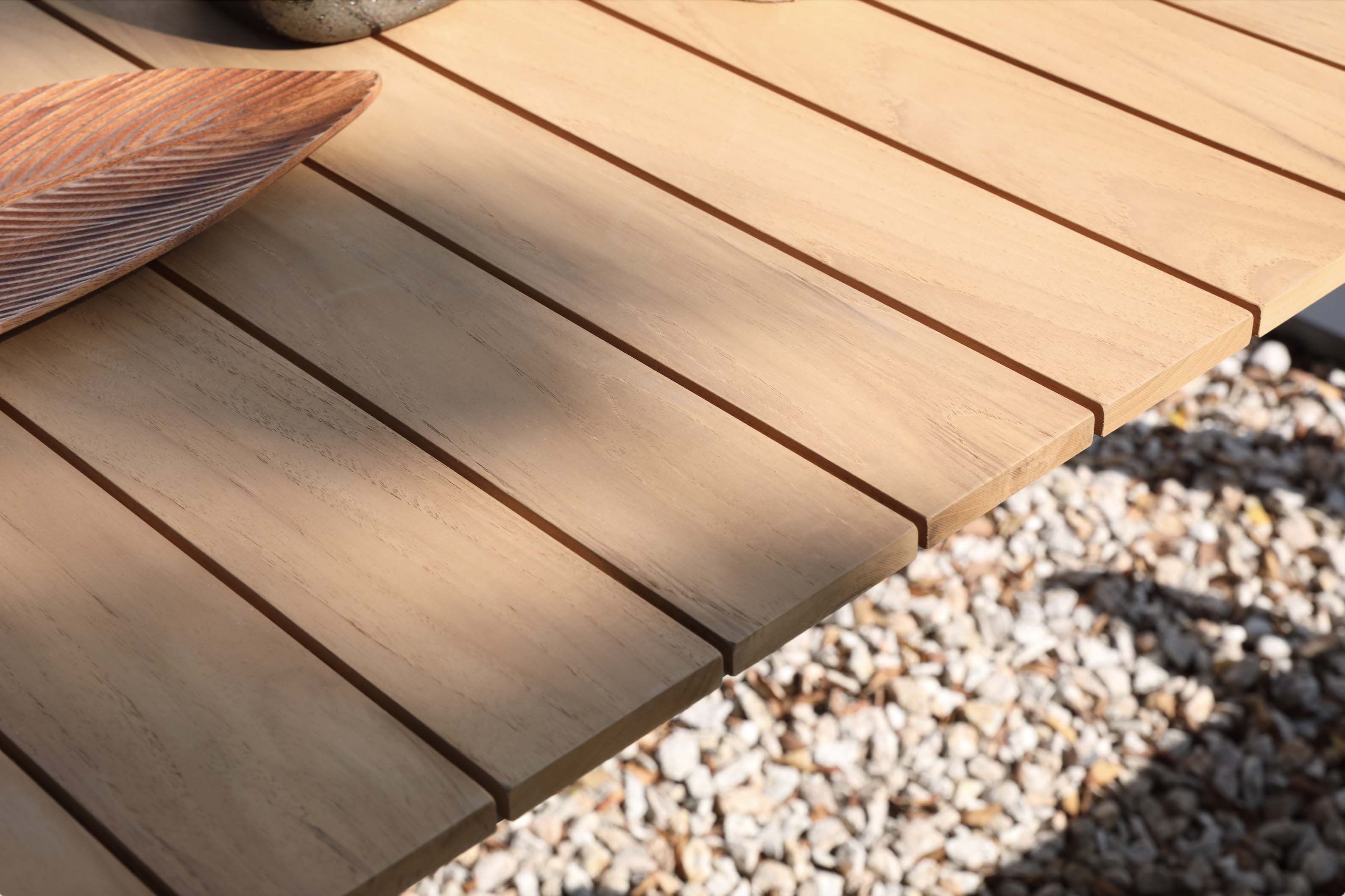 Plateau de table en teck FSC naturel aux lattes fines, posé sur sol en gravier, soulignant l'élégance méditerranéenne.