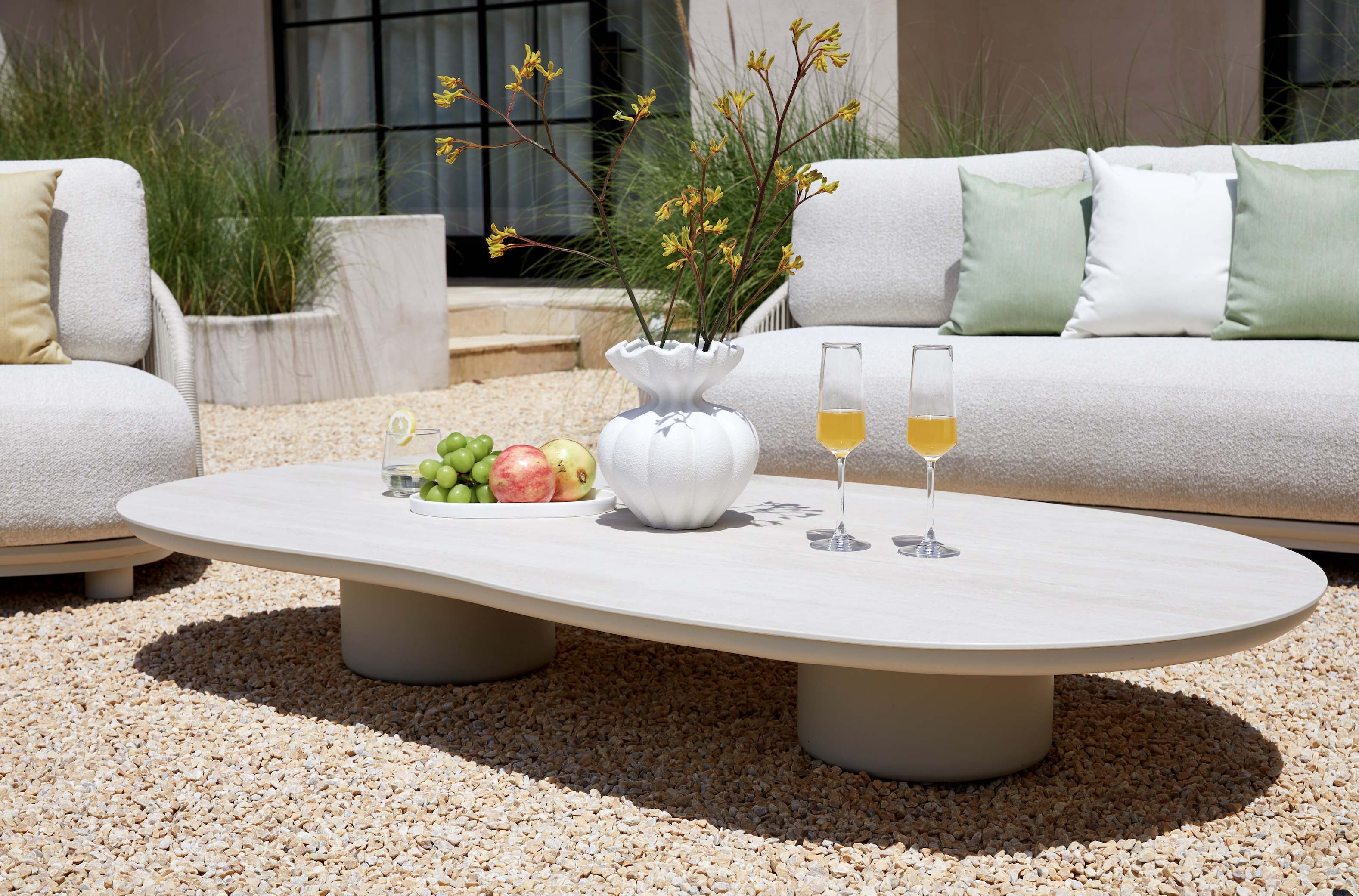 Table basse ovale en aluminium design avec plateau céramique beige, entourée de canapé en tissu 100% Oléfine Sunproof. 